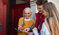 Eine ältere Frau an der Haustür im Gespräch mit zwei jungen Menschen. Sie schauen auf ein Klemmbrett. 