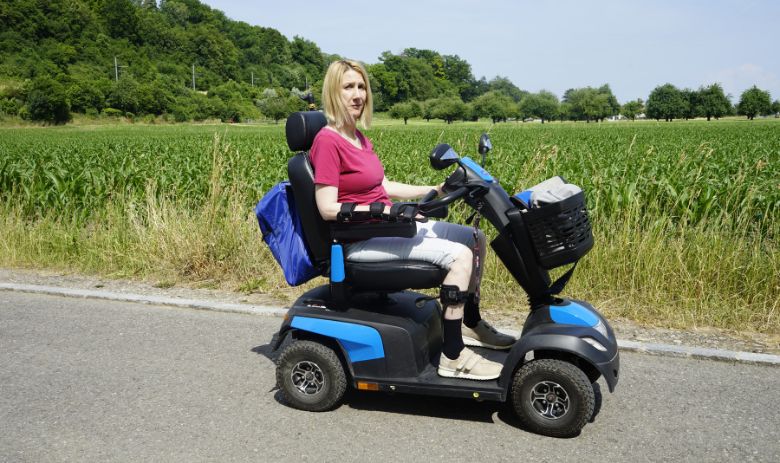 Une femme aux cheveux blonds est assise dans un fauteuil roulant électrique devant un champ à la végétation dense. L'expression de son visage est grave. 
