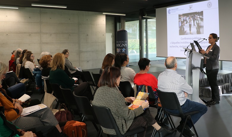 Une femme se tient derrière un pupitre, des personnes assises face à elle l'écoutent