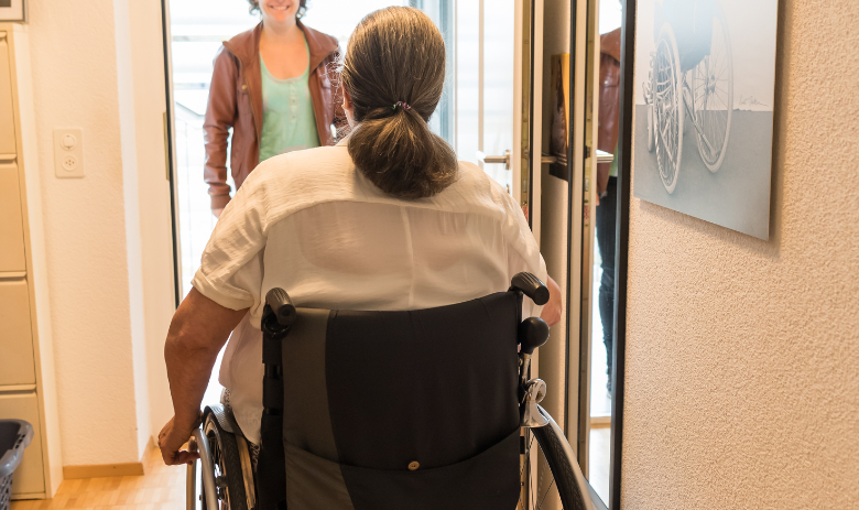 Services de transport Une femme en fauteuil roulant à la porte d'entrée de son appartement. Une femme attend dehors.