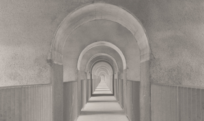 Ein schwarz-weiss-Foto eines langen Korridors. Auf beiden Seiten sind Geländer aus Metall, unterbrochen Wandabschnitten mit gewölbten Decken.